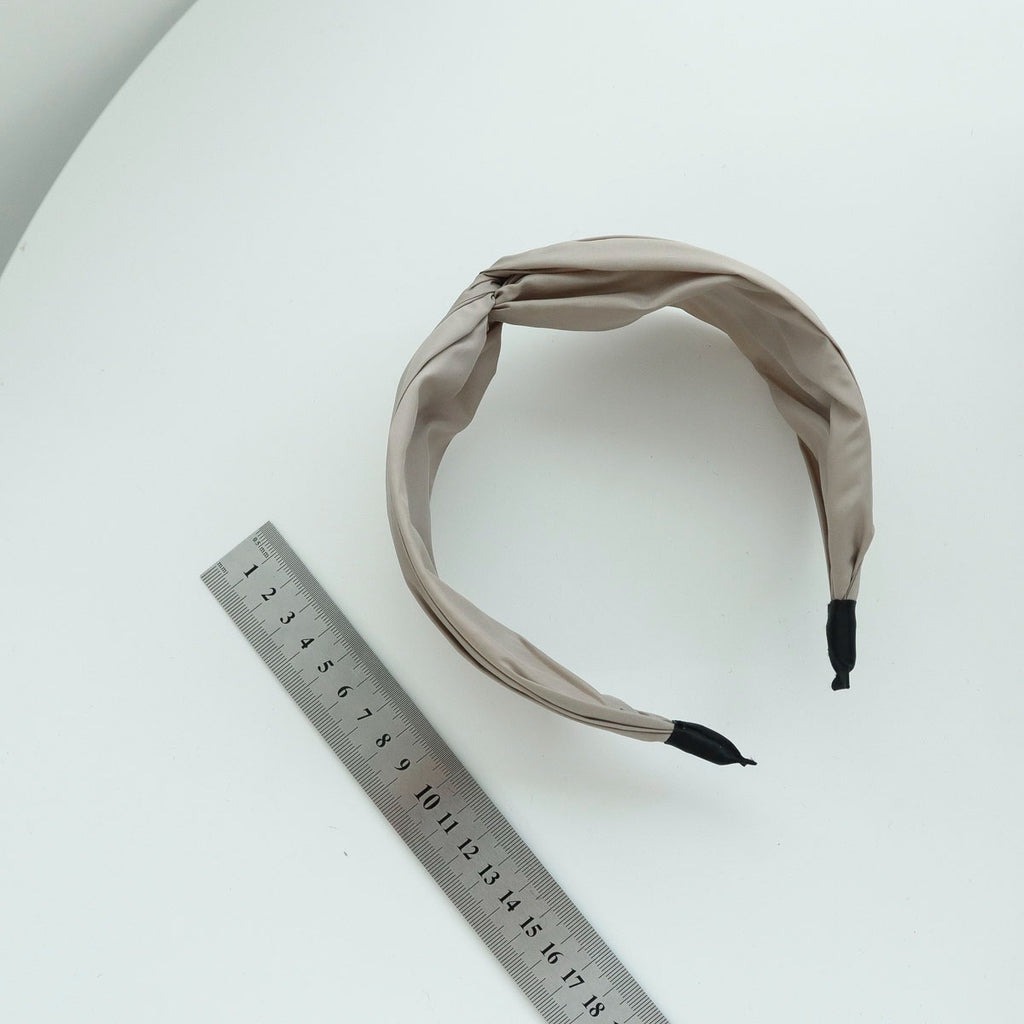 Top-down view of a beige wide knotted headband lying on a white table next to a standard metal measuring ruler to demonstrate product sizing.