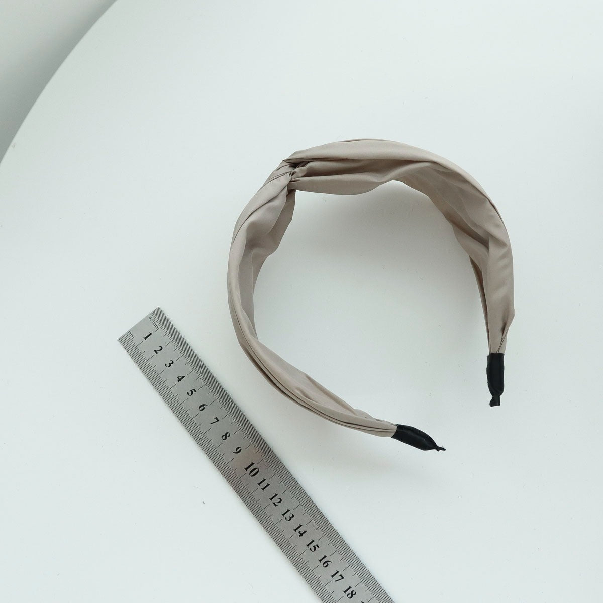 Top-down view of a beige wide knotted headband lying on a white table next to a standard metal measuring ruler to demonstrate product sizing.