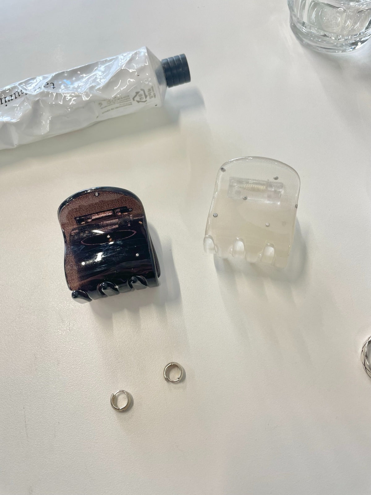 Close-up of a dark tortoiseshell brown and a milky ivory rhinestone studded mini hair claw clip sitting on a white desk next to two small silver hoop earrings.