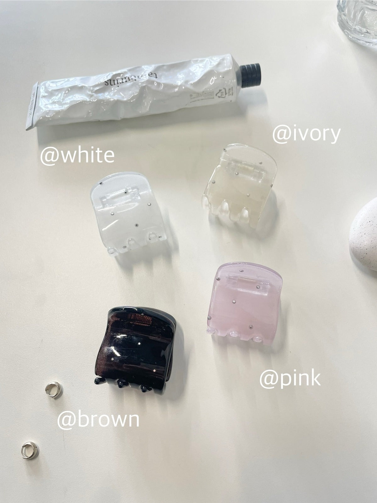 Top-down view of four rhinestone studded mini claw clips labeled @white, @ivory, @pink, and @brown, placed cleanly on a white surface with small silver hoop earrings.