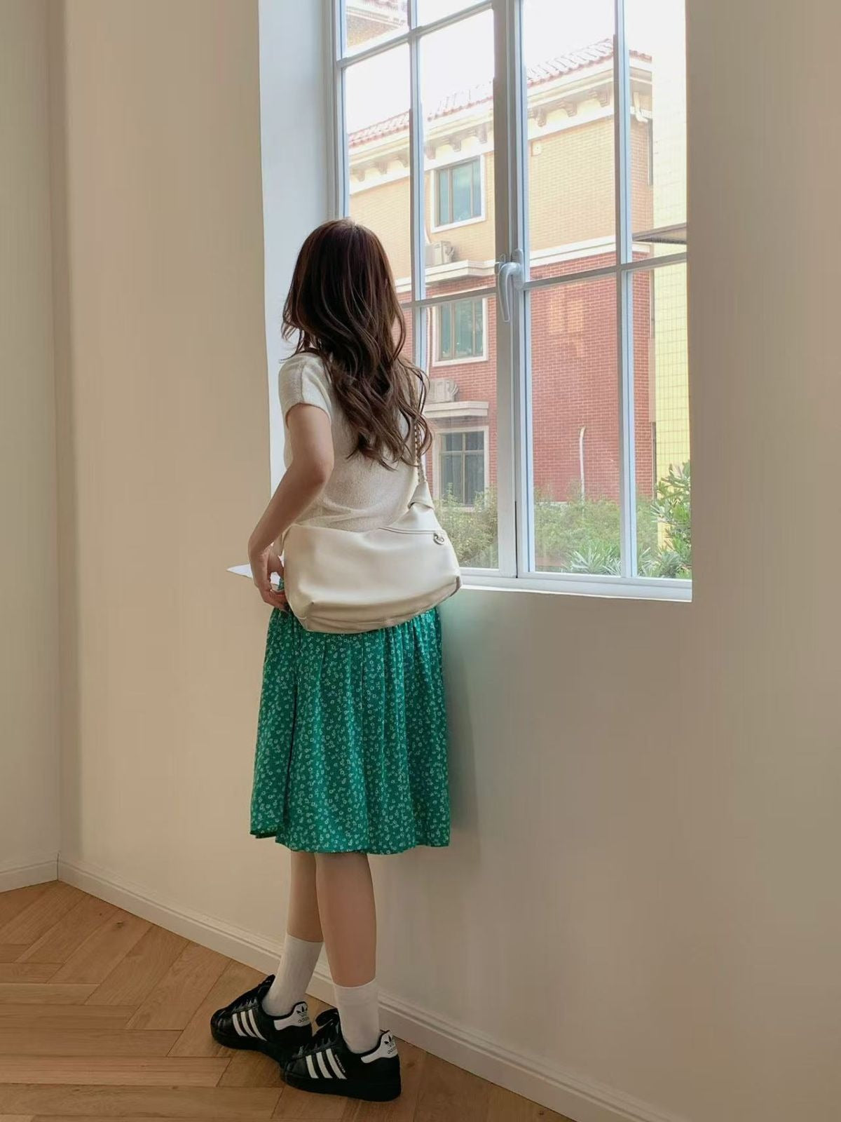 A woman looking out a window wearing a green floral skirt and carrying a cream white crescent shoulder bag with a braided strap detail.