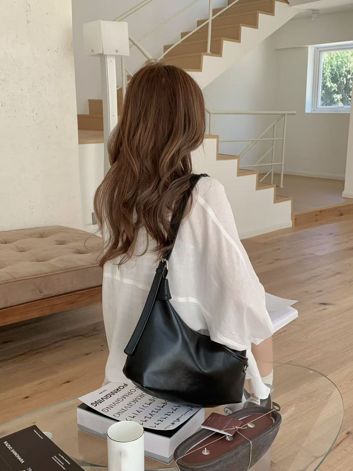 A woman in a white blouse sitting in a bright living room, wearing a black minimalist crossbody bag with an adjustable strap resting on her lap.