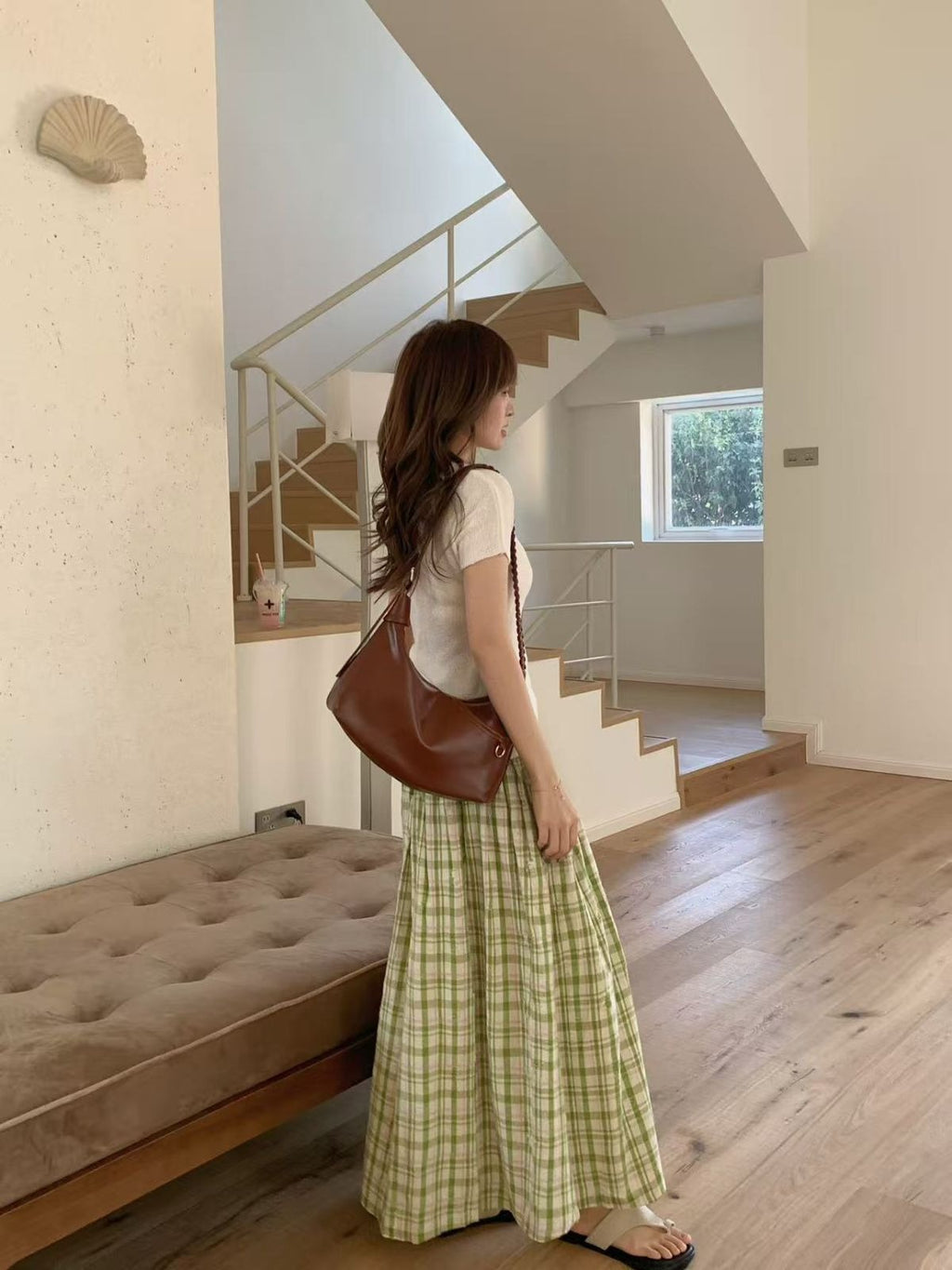 Side profile of a woman carrying a brown slouchy hobo bag on her shoulder, walking past a beige tufted bench in a minimalist house.