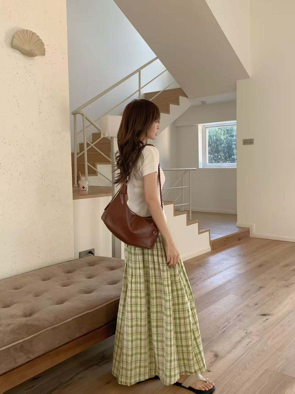 Side profile of a woman carrying a brown slouchy hobo bag on her shoulder, walking past a beige tufted bench in a minimalist house.
