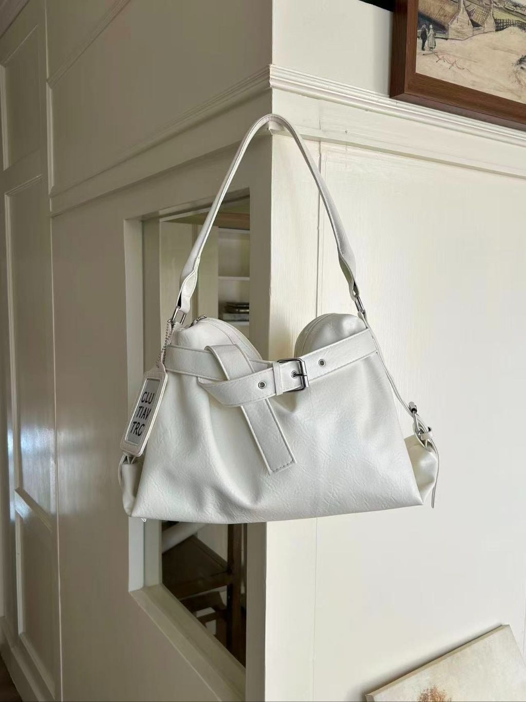 A cream white soft vegan leather hobo shoulder bag with a wide front belt strap hanging gracefully from a corner wall molding in a bright home.