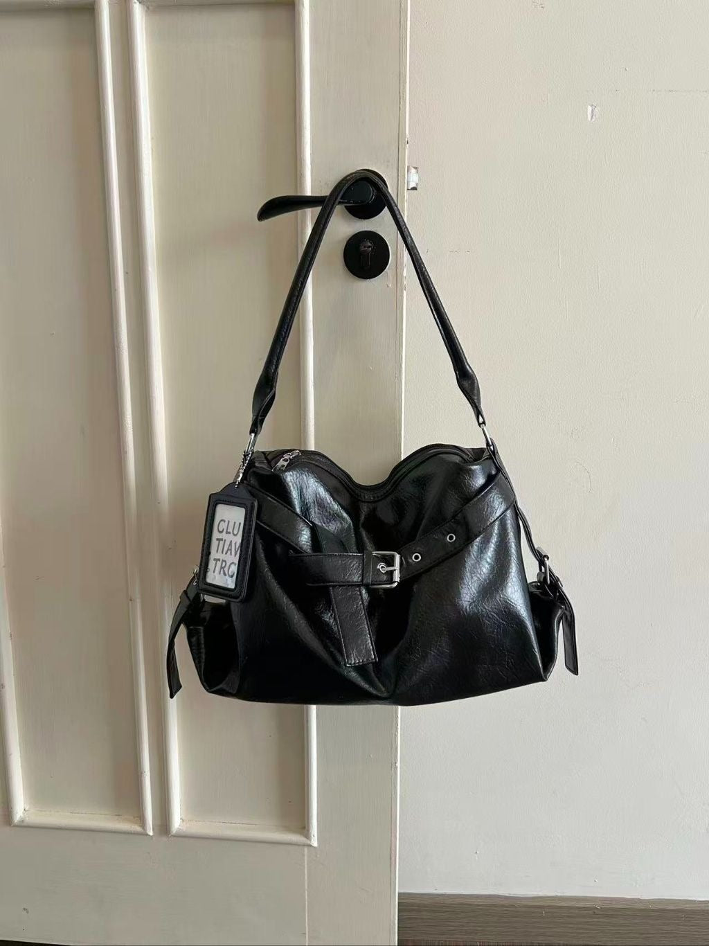 A black soft leather slouchy hobo bag with a front belt buckle strap and graphic tag hanging from a black door handle on a white door.