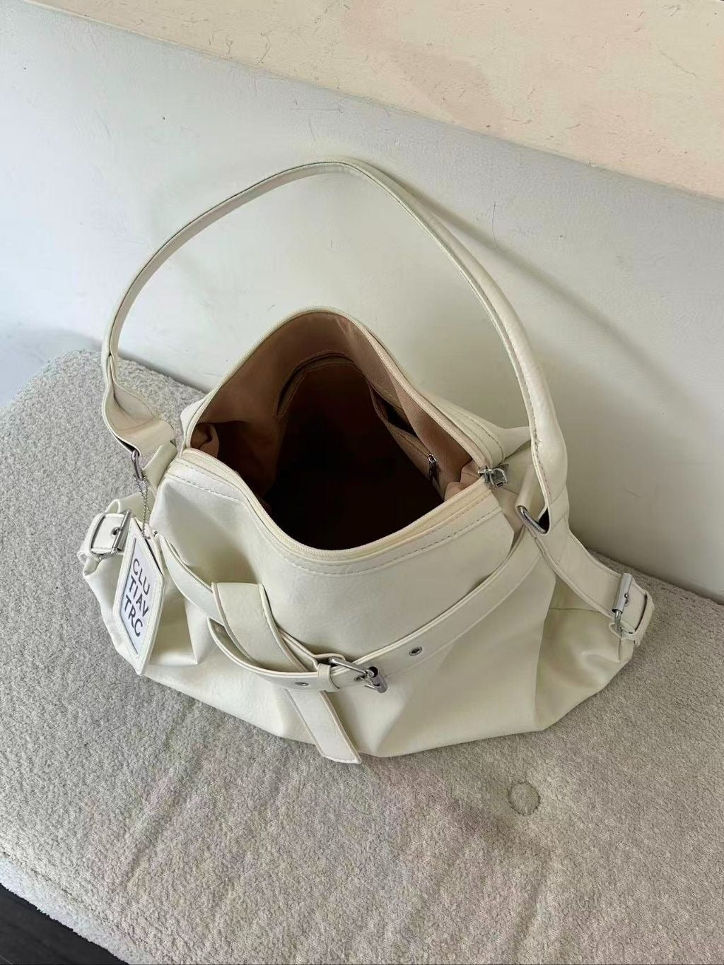 Top-down view into an open cream white slouchy hobo bag, revealing a roomy brown fabric-lined interior with zipper pockets, resting on a textured grey surface.