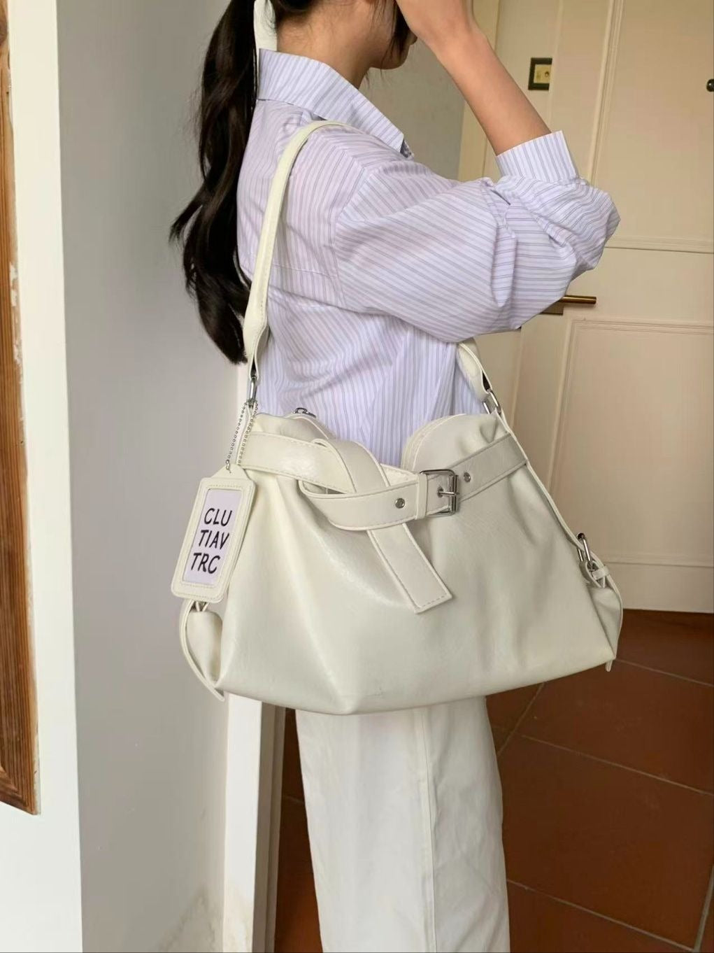 A woman in a striped shirt and white pants carrying a cream white slouchy hobo shoulder bag featuring a wide belt detail and graphic luggage tag.