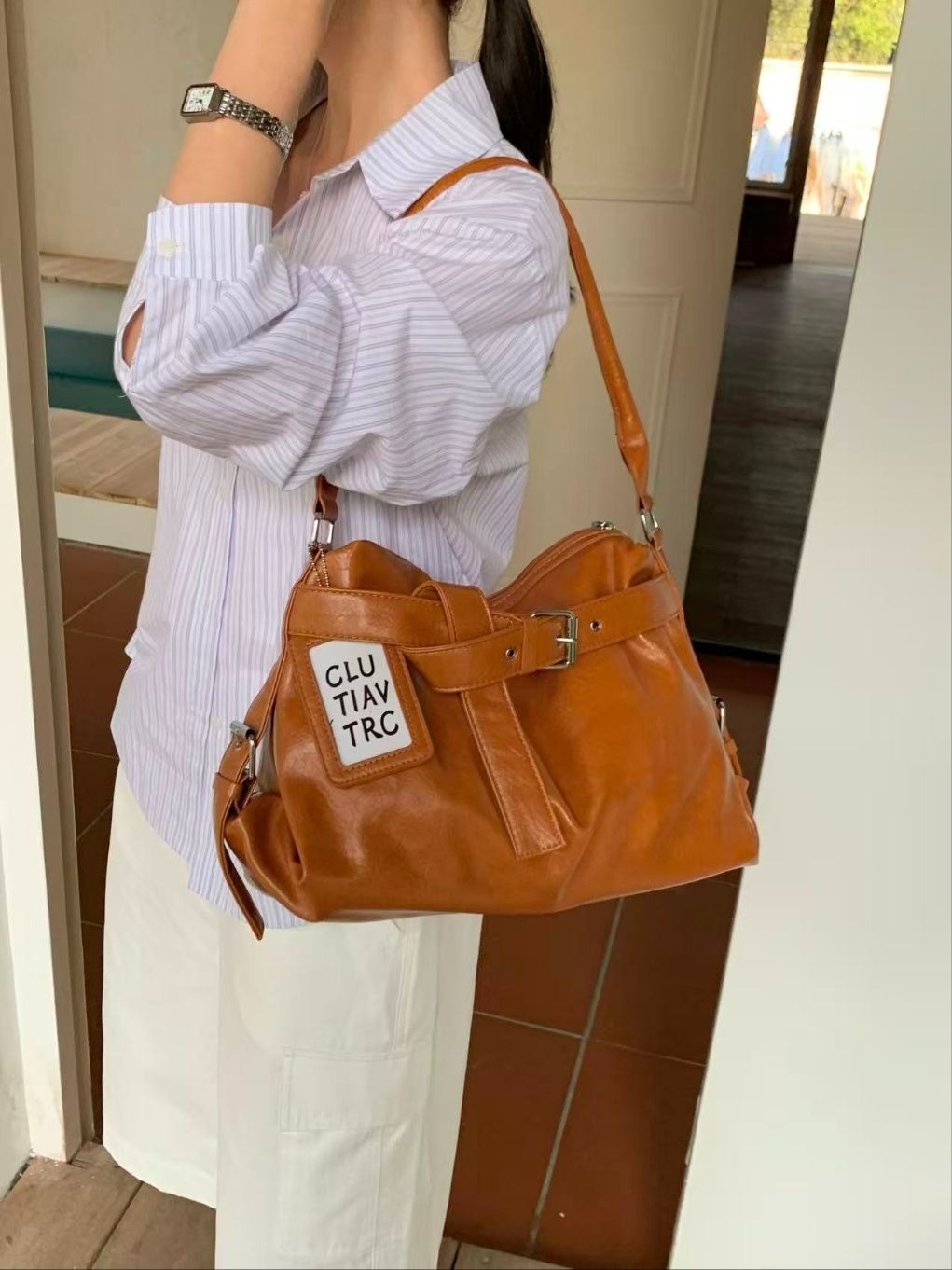 A woman taking a mirror selfie wearing a striped shirt, holding a caramel tan soft leather hobo bag with a belt strap detail on her shoulder.