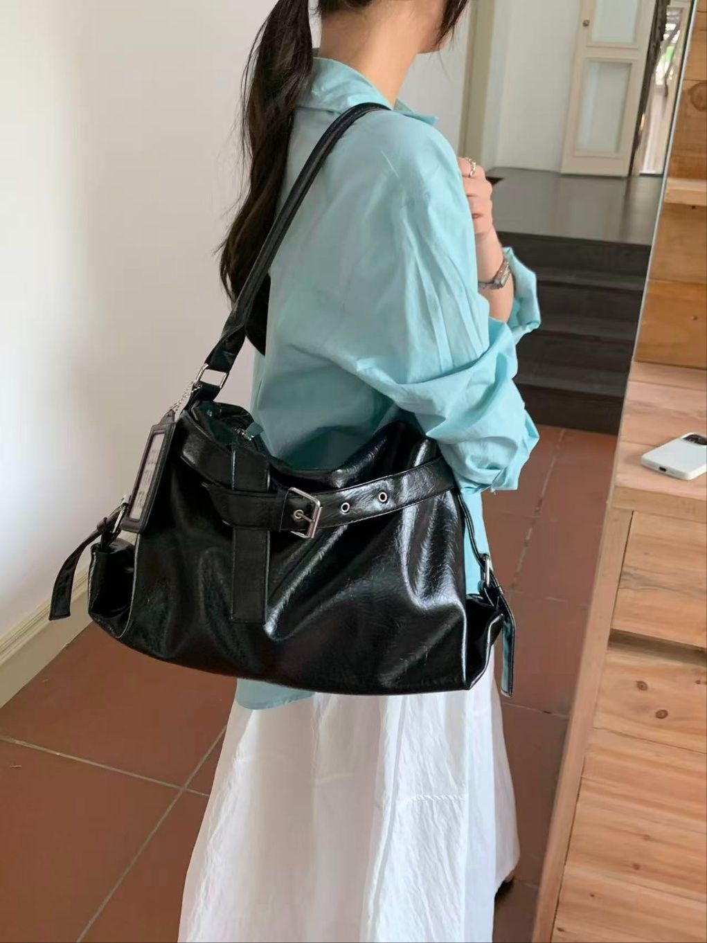 Back view of a woman with a low ponytail wearing a light blue shirt, carrying a black slouchy belt point hobo bag on her shoulder.