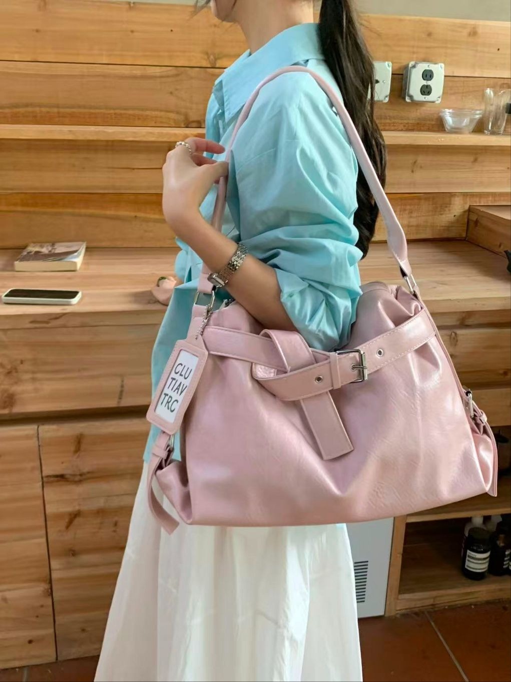A woman in a striped button-down shirt and white pants carrying a caramel tan slouchy hobo bag featuring a graphic tag and silver buckle hardware.