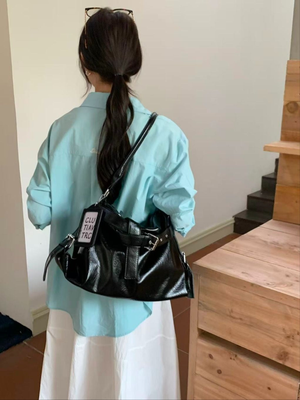 Side profile of a woman in a light blue top and white skirt carrying a black soft vegan leather shoulder hobo bag with a prominent front buckle strap.