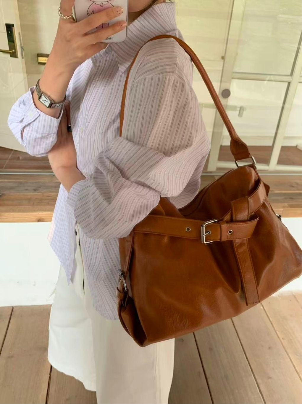 A woman wearing a light blue button-down shirt carrying a blush pink slouchy hobo bag with a front belt buckle detail and graphic luggage tag.