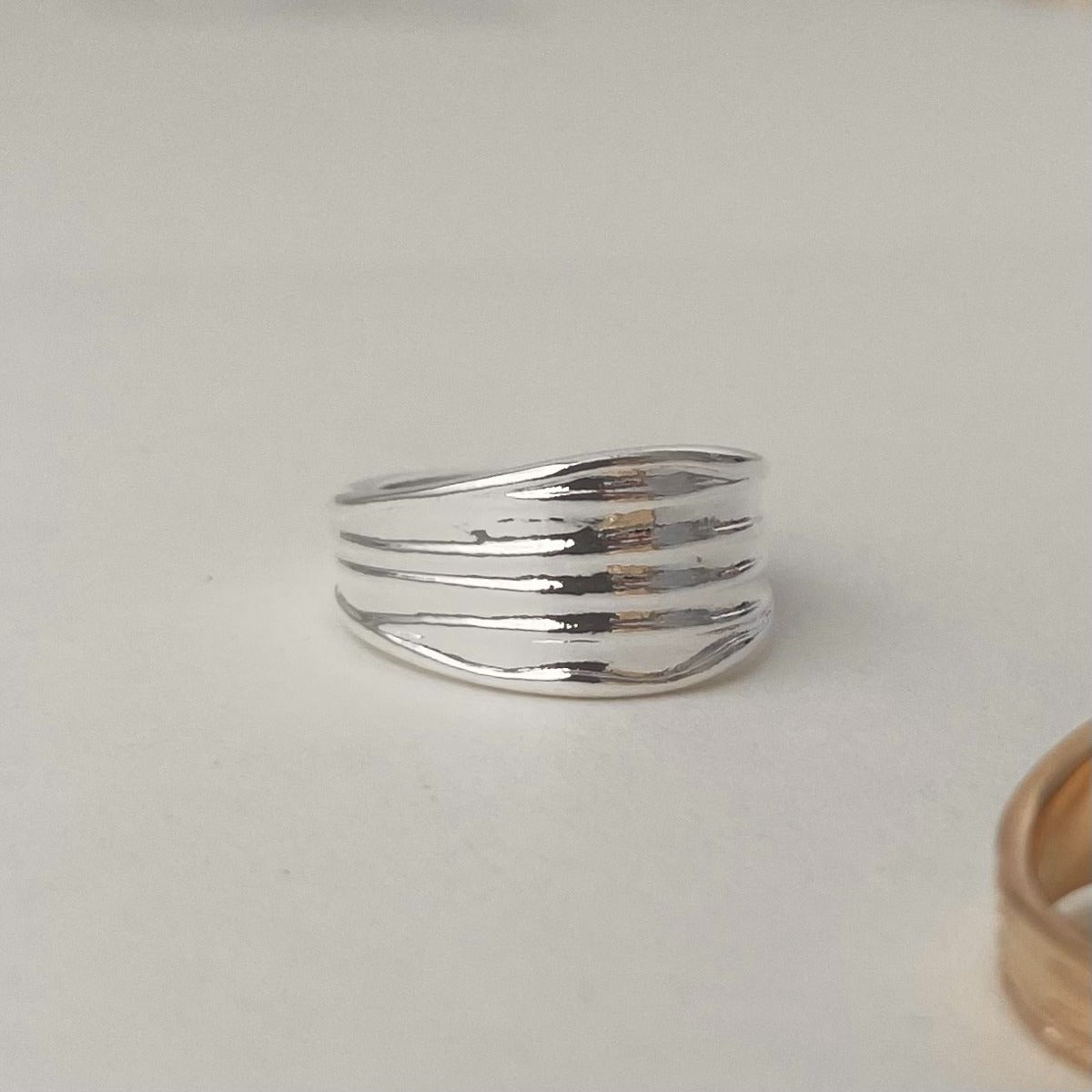 A shiny silver ridged wide band ring sitting next to a partially visible gold textured ring on a plain white background.