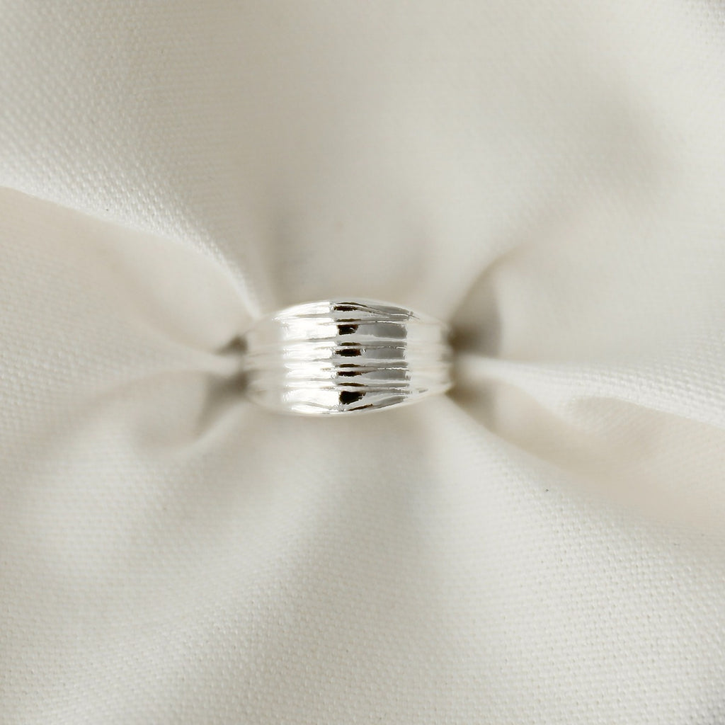 A sleek silver textured ribbed wide band ring resting on soft, gently folded white fabric.