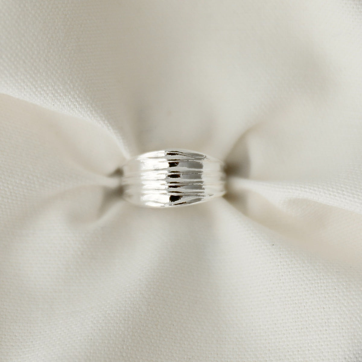 A sleek silver textured ribbed wide band ring resting on soft, gently folded white fabric.