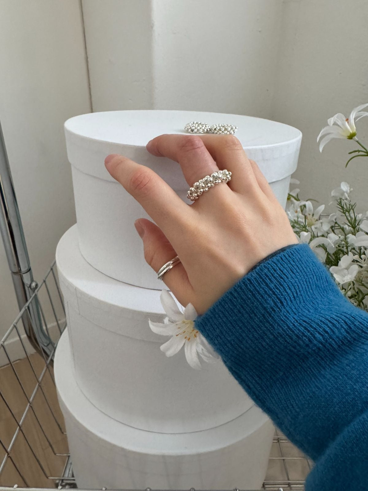 Hand wearing a cozy blue sweater and a chunky silver beaded elastic ring, resting gently on a stack of white circular hat boxes.
