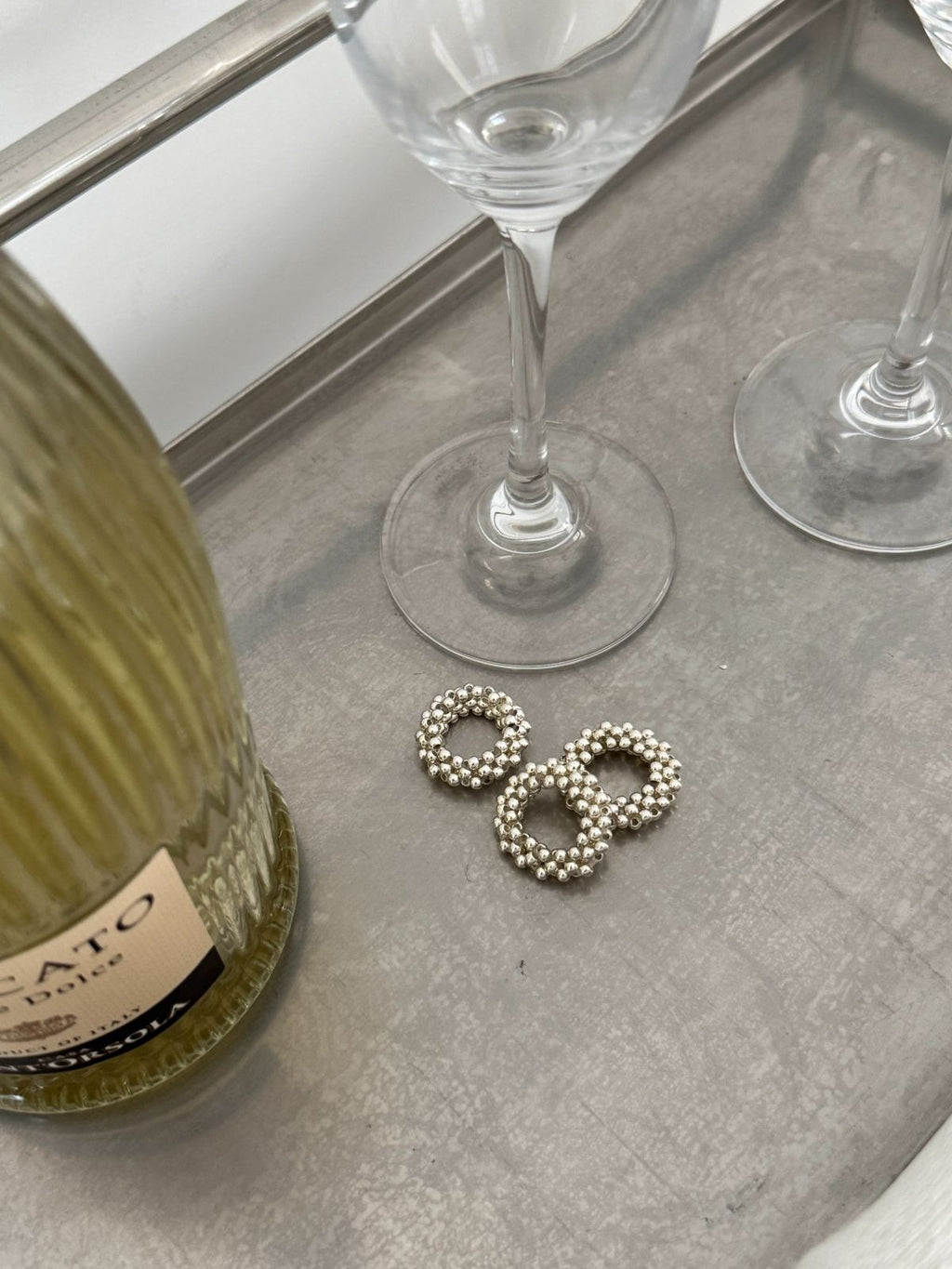 Three metallic silver stretch bead rings sitting on a textured grey serving tray next to the stems of empty wine glasses and a green glass bottle.
